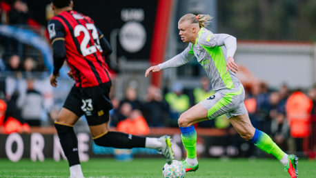 A soccer player in a grey and green kit is sprinting with the ball, being pursued by an opponent in a black and red kit on a field during a match.