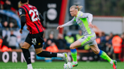 A soccer player in a grey and green kit is sprinting with the ball, being pursued by an opponent in a black and red kit on a field during a match.