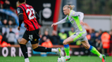 A soccer player in a grey and green kit is sprinting with the ball, being pursued by an opponent in a black and red kit on a field during a match.