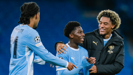 Three Manchester City players celebrating together on the field, wearing sky blue kits. One player is wearing a black jacket with the club logo.