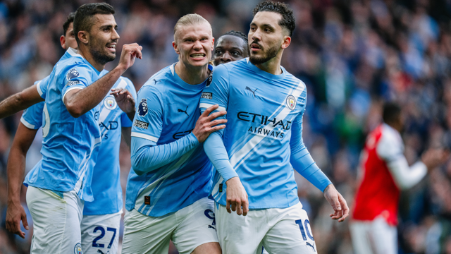 Manchester City players wearing blue jerseys with a sponsor logo celebrate a goal during a match. One player appears to be clapping.