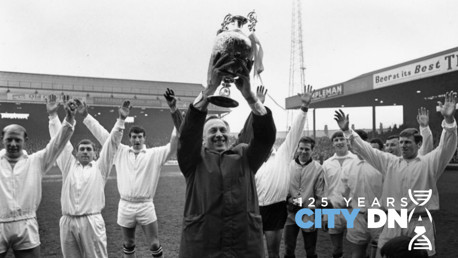 Black and white photo of a football team celebrating with a large trophy held aloft. Players are wearing white kits and raising their arms in celebration. The stadium backdrop features banners and a DNA symbol with '125 Years City DNA' text.