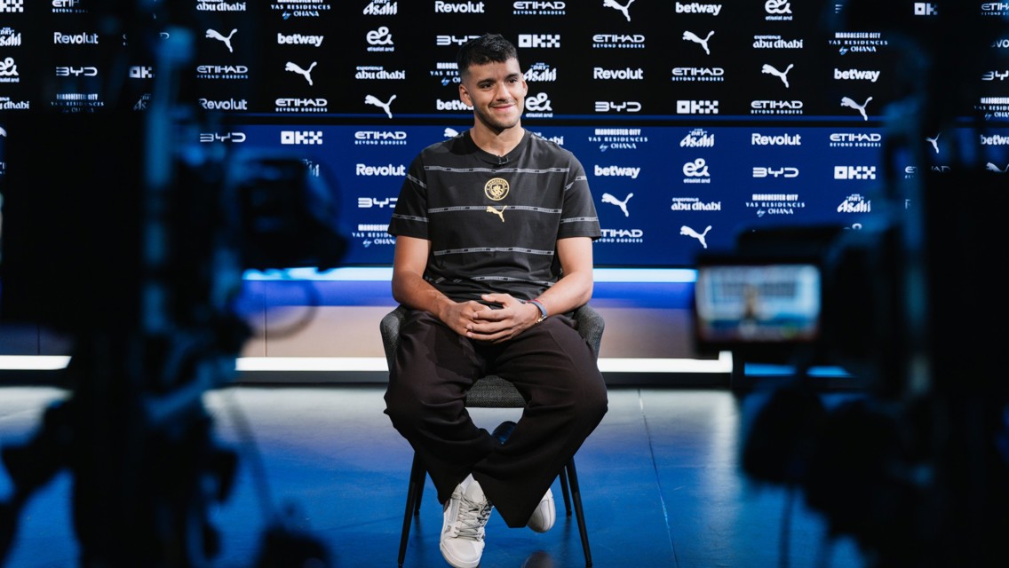 A person sitting in a media interview setup, wearing a black and gray striped shirt with Manchester City logos in the background.