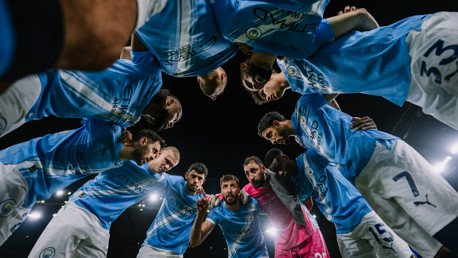 Soccer players in Manchester City kits form a huddle on the field, viewed from below. The lighting suggests a night match.