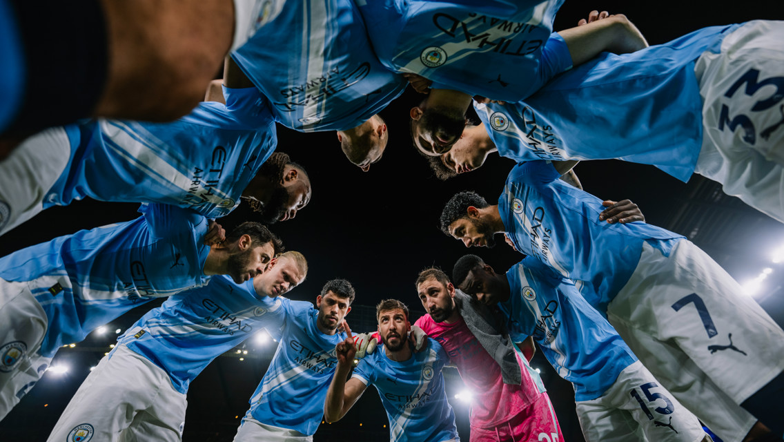 Soccer players in Manchester City kits form a huddle on the field, viewed from below. The lighting suggests a night match.
