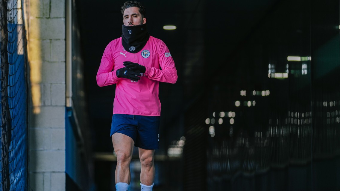 A soccer player wearing a pink Manchester City training jersey, gloves, and a neck warmer walks through a tunnel. The player's face is blurred.