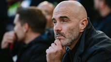 A bald man with a beard wearing a black jacket is thoughtfully observing something, his hand resting on his chin.
