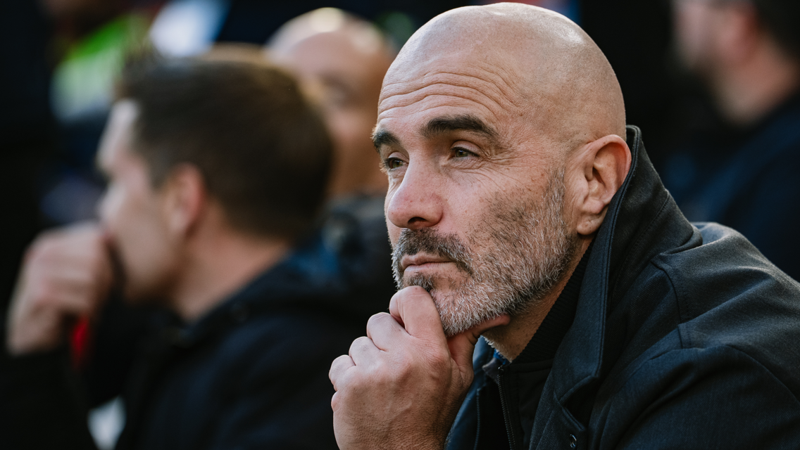 A bald man with a beard wearing a black jacket is thoughtfully observing something, his hand resting on his chin.