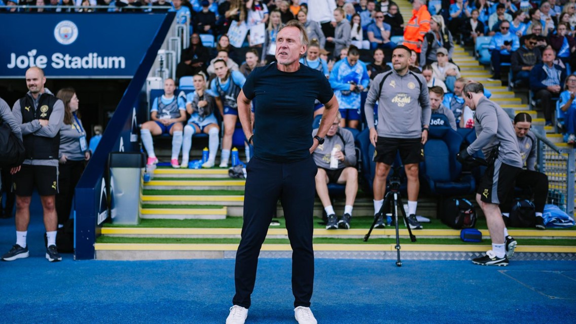A coaching staff member stands on the sideline at Joie Stadium, with Manchester City Women's players and staff visible on the bench and seats behind.