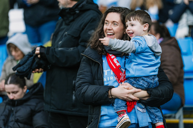A woman holding a child in the stands of Etihad Stadium, both wearing Manchester City gear and a scarf, surrounded by other fans.