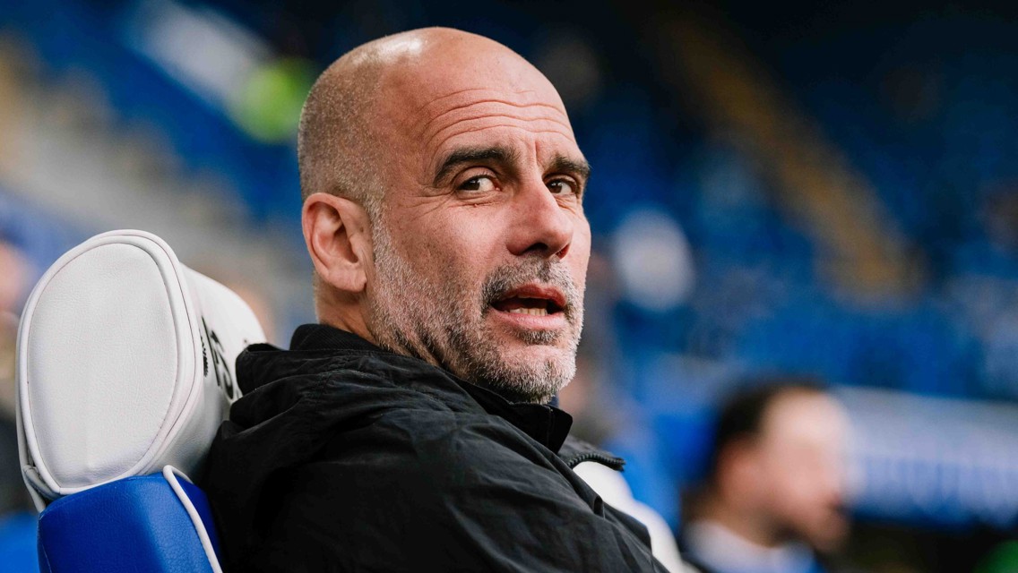 A person in a black jacket is seated on a white and blue chair in a stadium. The background is blurred with blue tones indicating a sports venue.