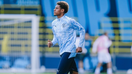 A young player in Manchester City attire is running on a football field during training. The backdrop shows goal posts and blurred figures.