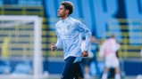 A young player in Manchester City attire is running on a football field during training. The backdrop shows goal posts and blurred figures.