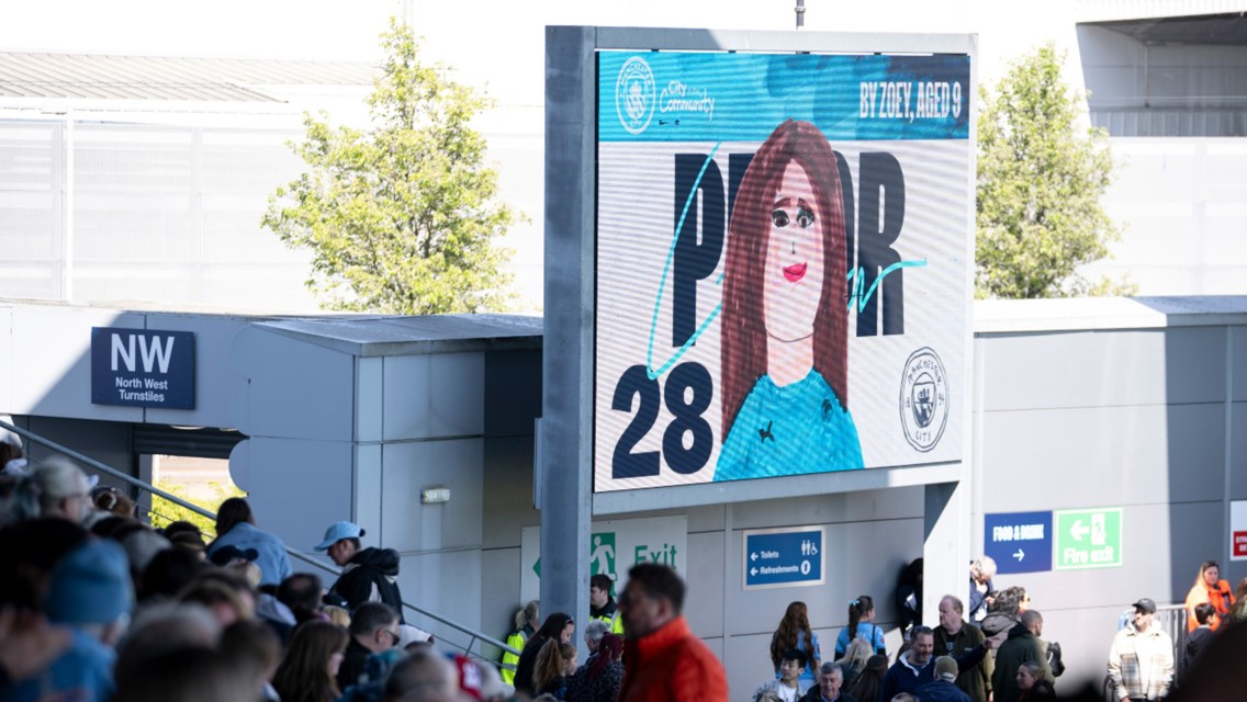 A large digital billboard at a stadium entrance displays a child's drawing of a soccer player with the message 'By Zoey, aged 9'.