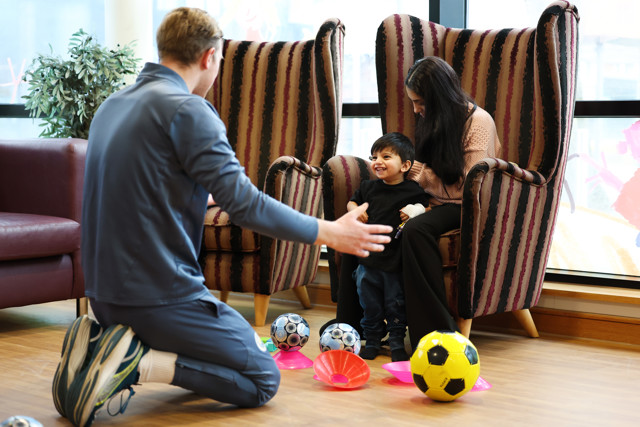 A coach is kneeling on the floor, engaging with a child seated on a woman's lap in a room scattered with colorful footballs and cones.