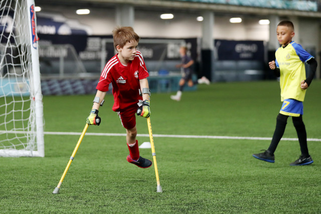 A young boy on crutches wearing a red soccer uniform plays on a football field, with another boy in a yellow uniform running in the background.