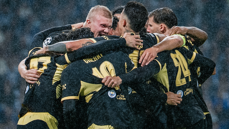 A group of Manchester City players celebrating a goal during a match, wearing black and yellow kits with visible water droplets, showcasing team unity and excitement.