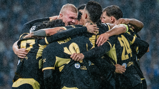 A group of Manchester City players celebrating a goal during a match, wearing black and yellow kits with visible water droplets, showcasing team unity and excitement.