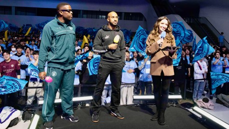A Manchester City fan event featuring two people on stage wearing Manchester City attire with microphones, and a female presenter holding a microphone and a tablet. Fans in the background are waving Manchester City flags.