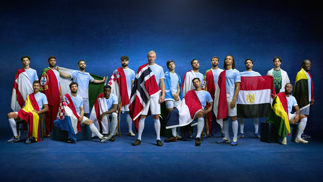 Group of 16 people wearing Manchester City kits, each with a different national flag draped over their shoulders, standing against a blue background.