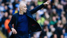 A bald man wearing a dark suit and blue shirt is pointing his finger while standing outdoors, with a blurred crowd in the background.