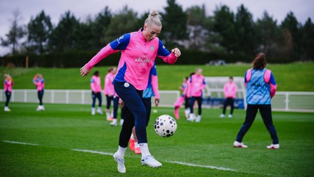 A football player from a women's team is skillfully controlling a ball during a training session on a green field. Other players are visible in the background.