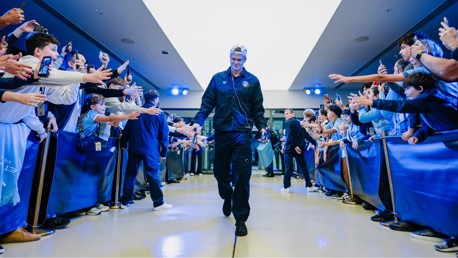A football player is walking through a corridor, high-fiving fans lined up on both sides. The atmosphere is celebratory with people reaching out and taking photos.