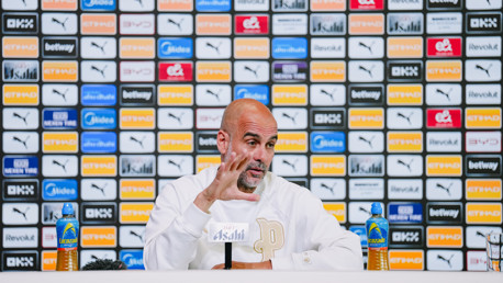 A person at a press conference with a Manchester City backdrop featuring multiple sponsor logos, a mic, and two bottles of Lucozade on the table.