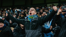 A crowd of Manchester City fans celebrate at the Etihad Stadium, wearing team scarves and cheering, with some recording on their phones. The front fan wears a Berghaus jacket.