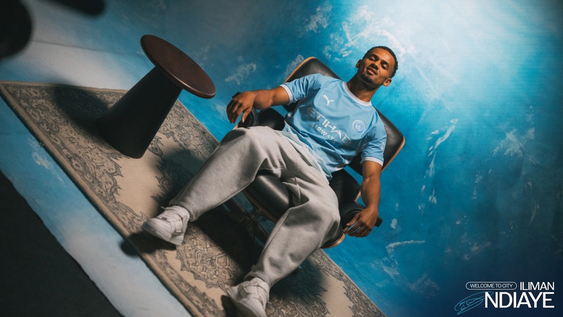 A person in a Manchester City football jersey is sitting on a chair against a blue background with text saying 'Welcome to City Iliman Ndiaye.'