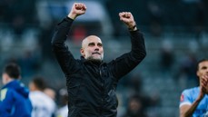A football coach wearing a black jacket celebrates with fists raised. The background shows blurred figures and a sports field.