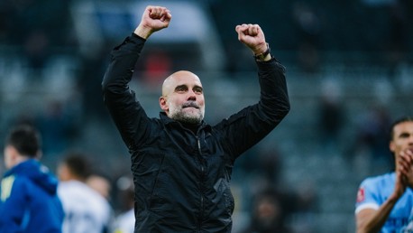 A football coach wearing a black jacket celebrates with fists raised. The background shows blurred figures and a sports field.