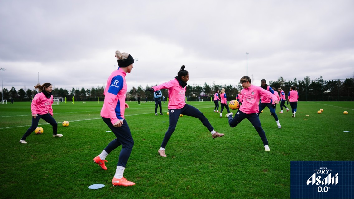 Players wearing pink training jerseys practice on a grassy field, interacting with footballs. Asahi logo is visible in the corner.
