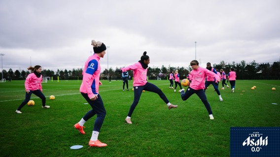 Players wearing pink training jerseys practice on a grassy field, interacting with footballs. Asahi logo is visible in the corner.