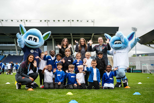 Group photo of children, coaches, and two Manchester City mascots at Academy Stadium during a community event.