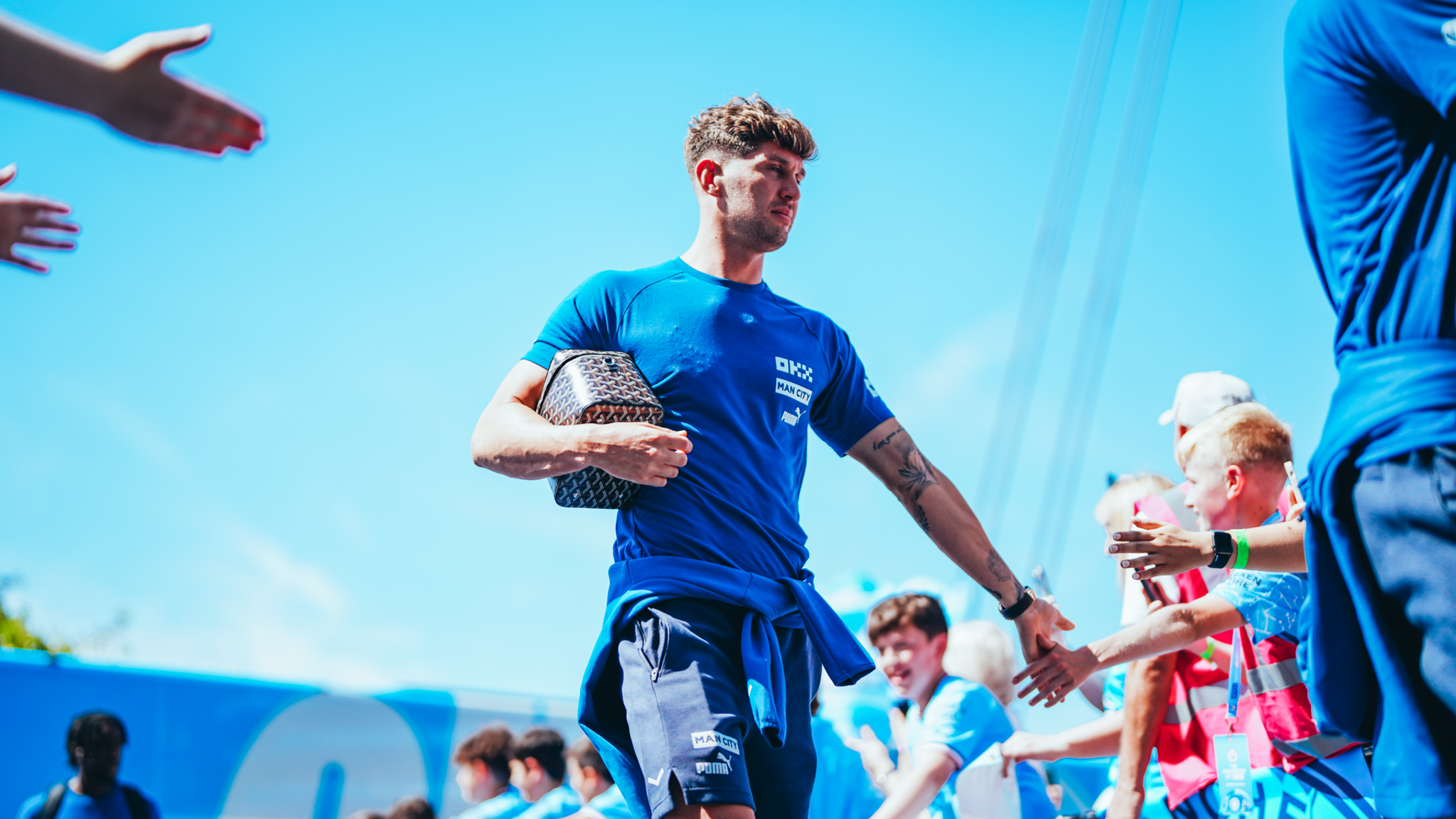 A Manchester City player high-fives fans while holding a designer bag, wearing a blue Man City T-shirt against a clear blue sky.