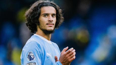 A Manchester City player wearing the team's kit, with the Premier League logo on the sleeve, clapping and facing the camera.
