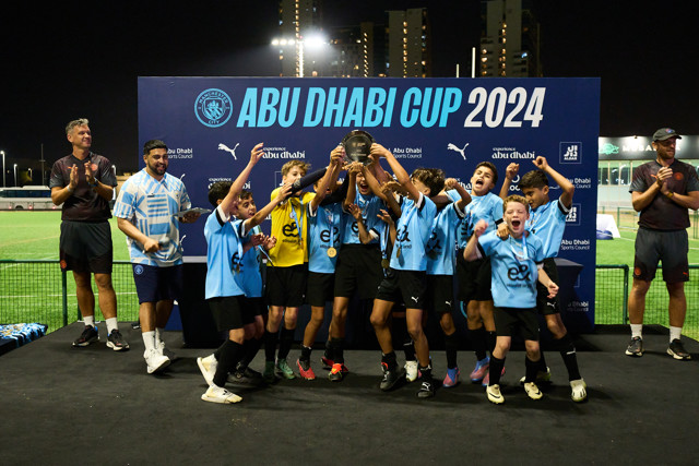 A youth football team is celebrating on stage holding a trophy at the Abu Dhabi Cup 2024. They are wearing blue shirts and have medals around their necks. The backdrop has the event title and sponsors like Manchester City and Puma.