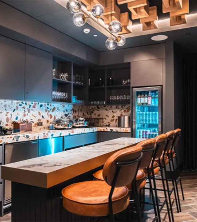 A modern kitchen with a long counter and four stools, featuring stylish lighting, shelving with glassware and a fridge filled with drinks.