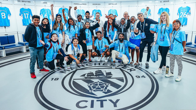 A group of people in a Manchester City locker room with jerseys displayed, gathered around the team's logo on the floor, wearing Manchester City gear and credentials.