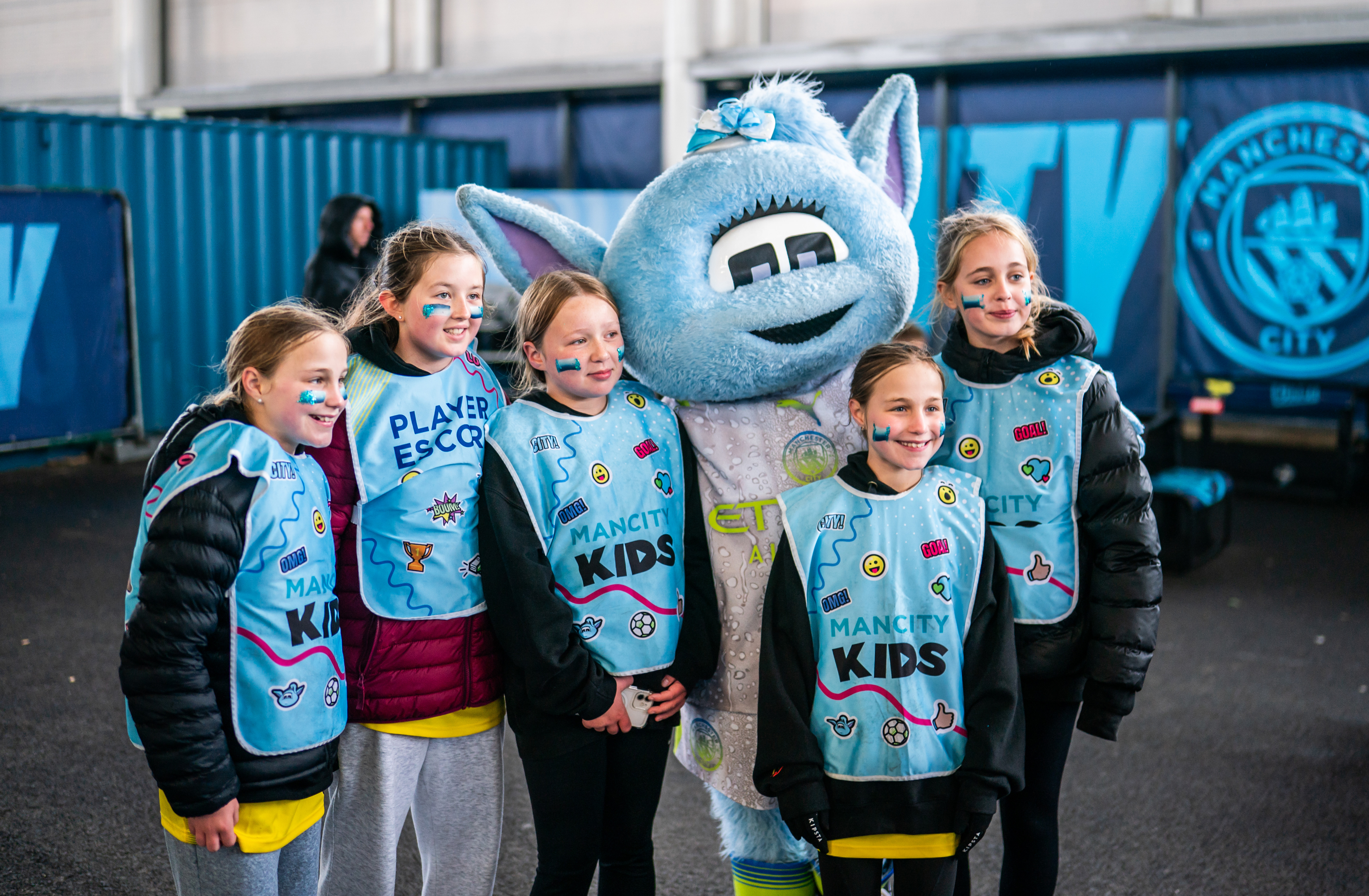 Group of children with the Manchester City mascot, wearing 'Man City Kids' bibs at an indoor space with Manchester City branding.