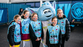 Group of children with the Manchester City mascot, wearing 'Man City Kids' bibs at an indoor space with Manchester City branding.