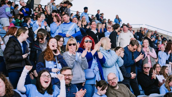 A crowd of fans wearing Manchester City jerseys and scarves is cheering and applauding in a stadium setting.