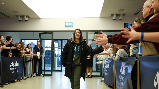 A person in casual attire walks through a tunnel while people on both sides reach out to greet them. The tunnel has blue branded banners and is well-lit.