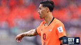 Soccer player in an orange jersey with the Netherlands flag, wearing number 11, pointing towards the side. The player has tattoos on their left arm. In the background, there is a blurred crowd in red.