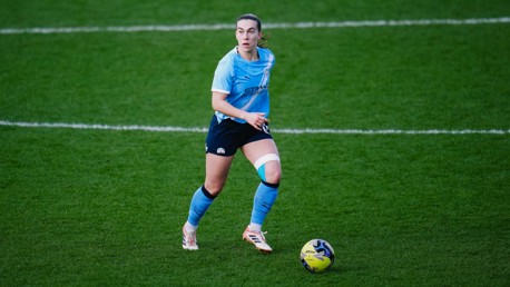 A female soccer player wearing a light blue Manchester City kit is on the field controlling a ball during a match.