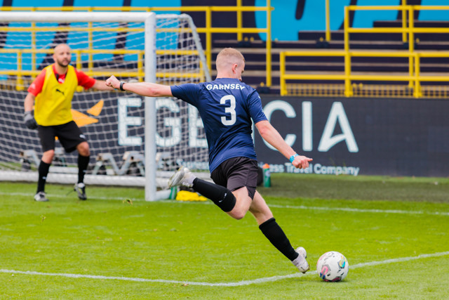 A soccer player, wearing a blue jersey with the name 'GARNSEY' and number '3', kicks the ball towards the goal, with the goalkeeper in yellow attempting to block.