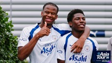 Two Manchester City players in a white and blue jersey smile and pose for a photo, one giving a thumbs-up. 'Asia Tour '26 presented by Abu Dhabi' is visible on the bottom right.