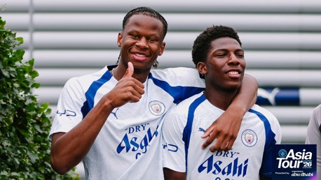 Two Manchester City players in a white and blue jersey smile and pose for a photo, one giving a thumbs-up. 'Asia Tour '26 presented by Abu Dhabi' is visible on the bottom right.
