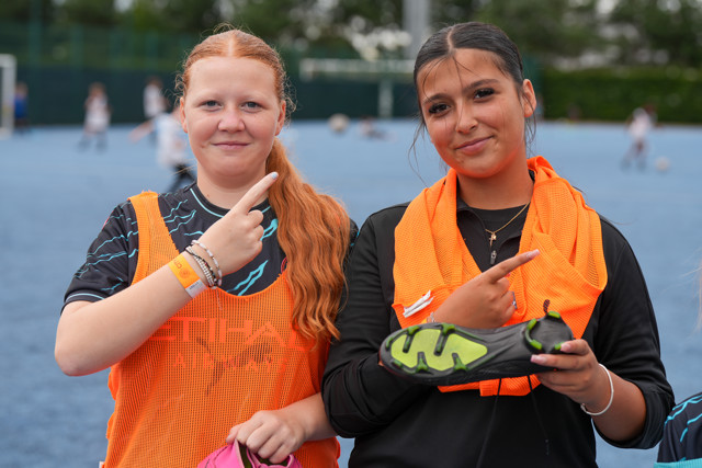 Two young female footballers with blurred faces, wearing Manchester City training gear. One is holding a football cleat, both pointing with their other hand. The background shows an outdoor football pitch.